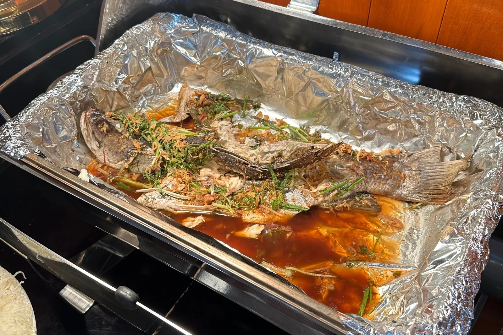 A whole cooked fish topped with herbs and fried shallots in a foil-lined steel chafing dish on a buffet counter, with varnished wood paneling behind it and a plate of noodles at the left edge.