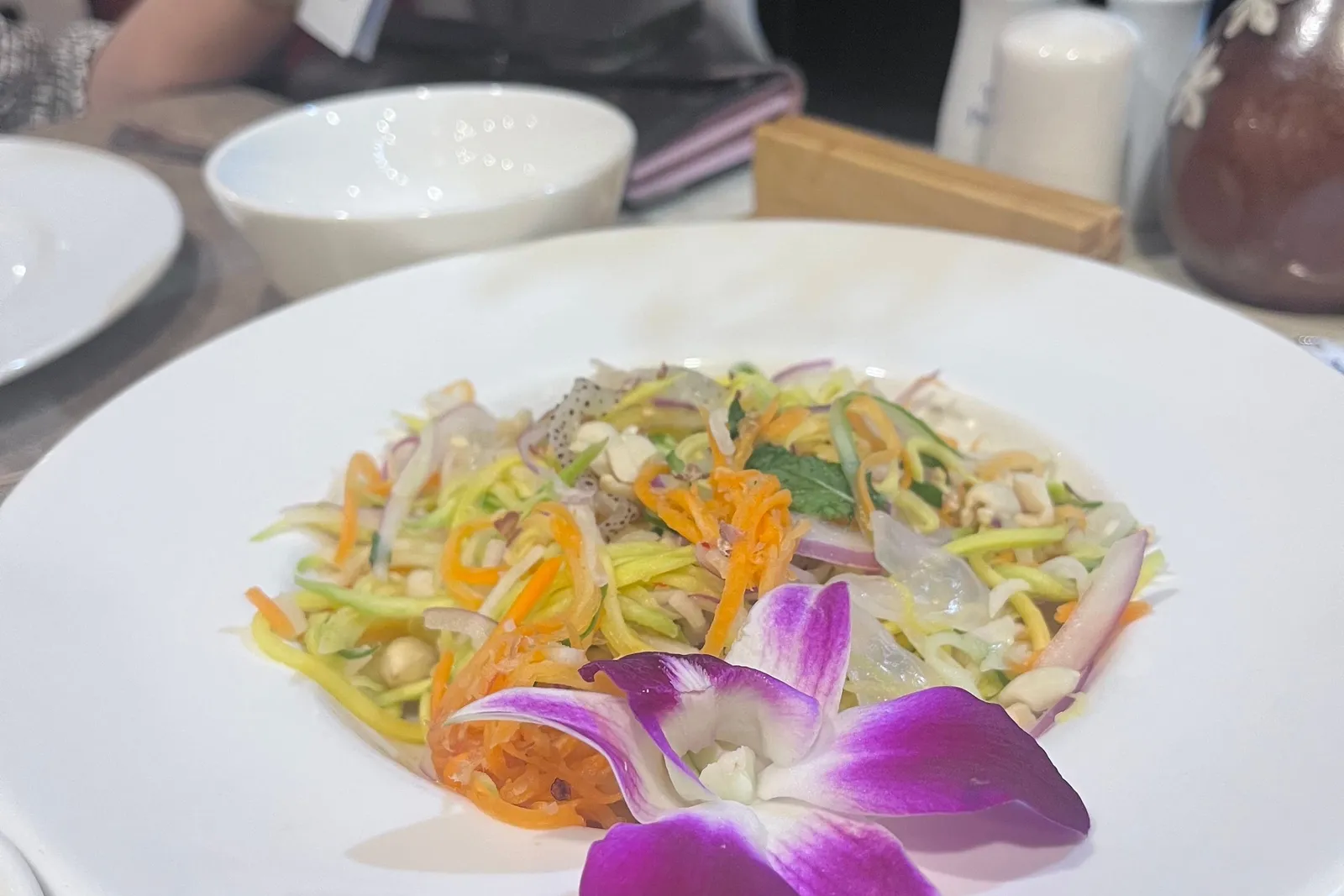 A white plate of shredded green mango and carrot salad topped with a purple orchid, on a dining table with a person holding a menu, salt and pepper shakers, and a vase in the background.