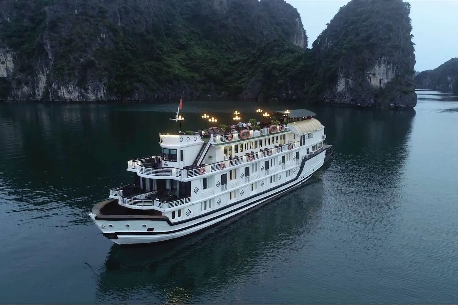 An aerial dusk view of a white multi-deck cruise boat with lit lamps on its upper deck, sailing on calm water beside forested limestone karst cliffs.