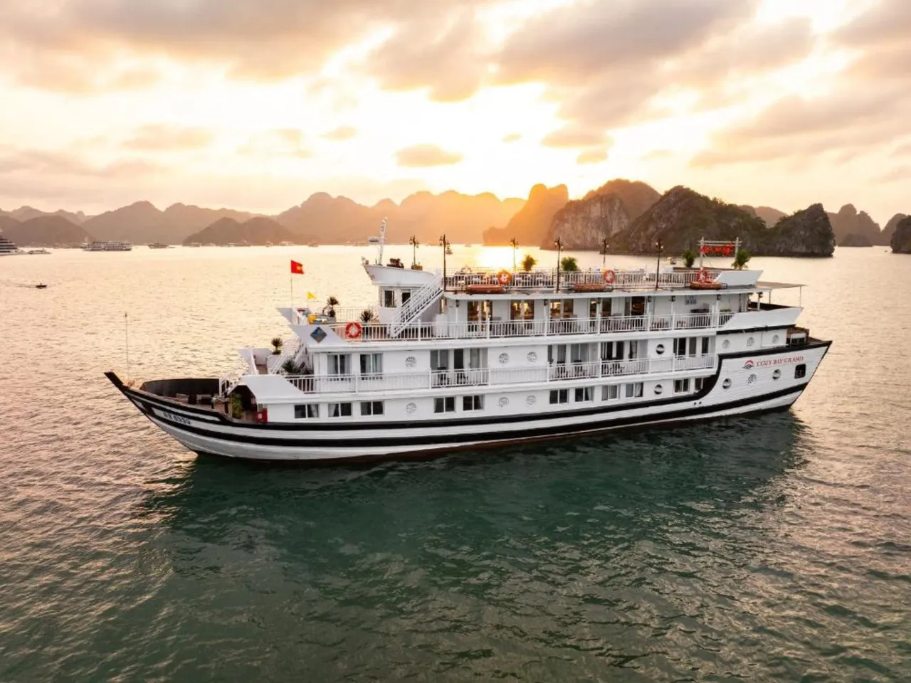 An aerial view of a white multi-deck cruise boat with a Vietnamese flag and hull branding sailing on calm water toward limestone karsts under a golden sunset sky.