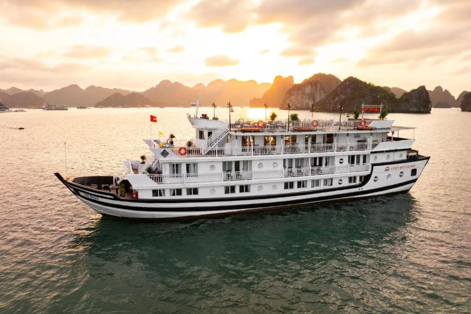 An aerial view of a white multi-deck cruise boat with a Vietnamese flag and hull branding sailing on calm water toward limestone karsts under a golden sunset sky.