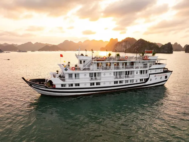 An aerial view of a white multi-deck cruise boat with a Vietnamese flag and hull branding sailing on calm water toward limestone karsts under a golden sunset sky.