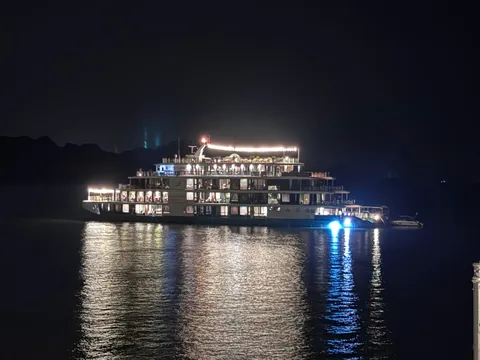 A multi-deck cruise ship lit up at night on dark water, with a small boat beside it, dark hills in the background, and a railing post at the right edge of the frame.