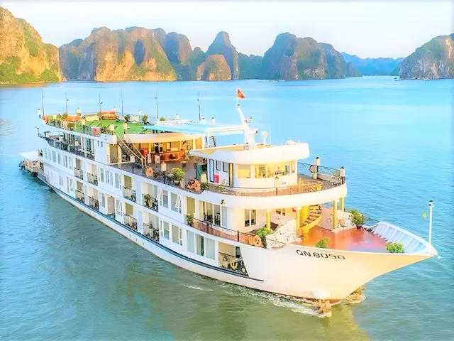 An aerial view of a multi-deck white cruise ship with the registration 'QN 8050' on its bow, sailing on calm turquoise water in front of limestone karst islands.