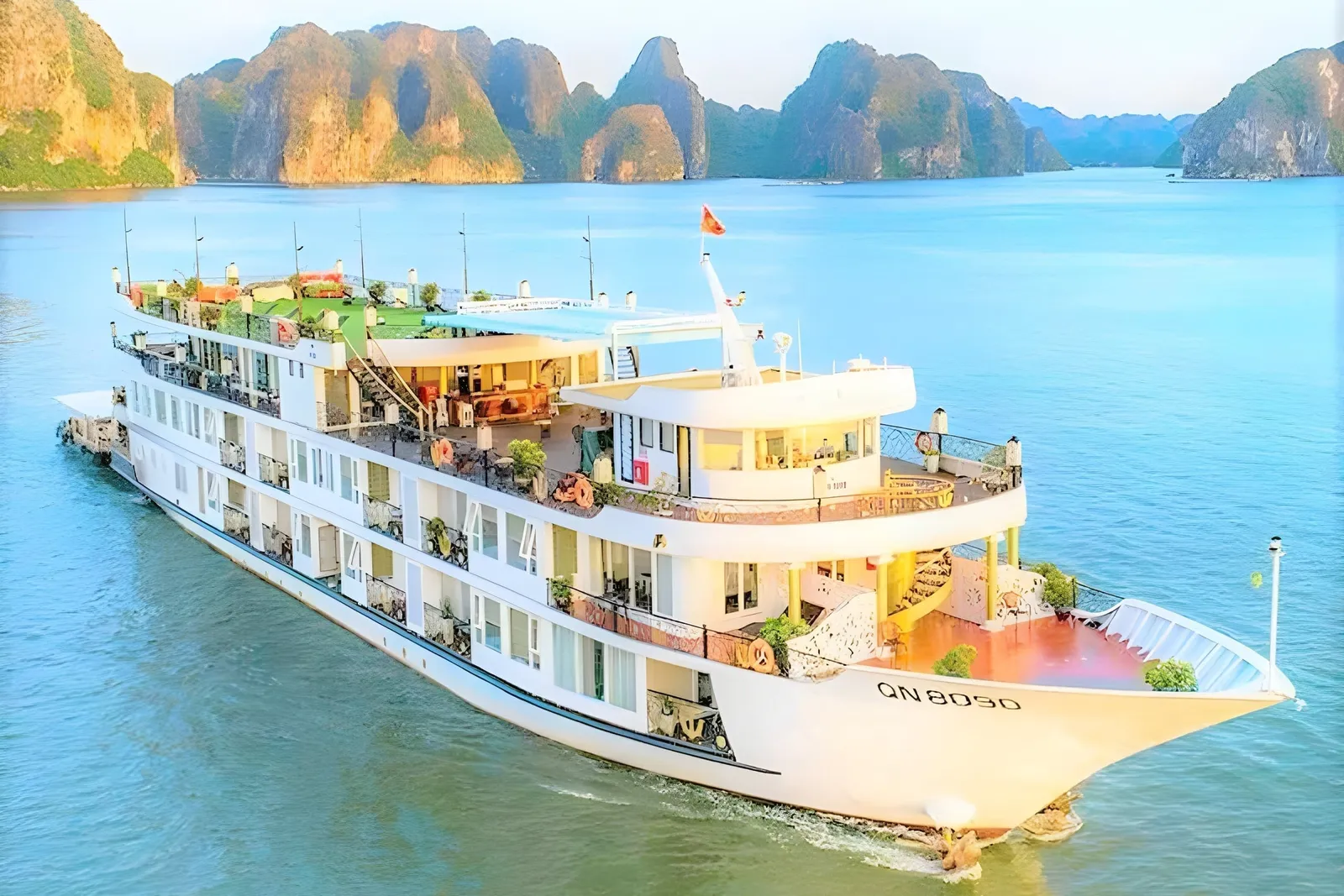 An aerial view of a multi-deck white cruise ship with the registration 'QN 8050' on its bow, sailing on calm turquoise water in front of limestone karst islands.