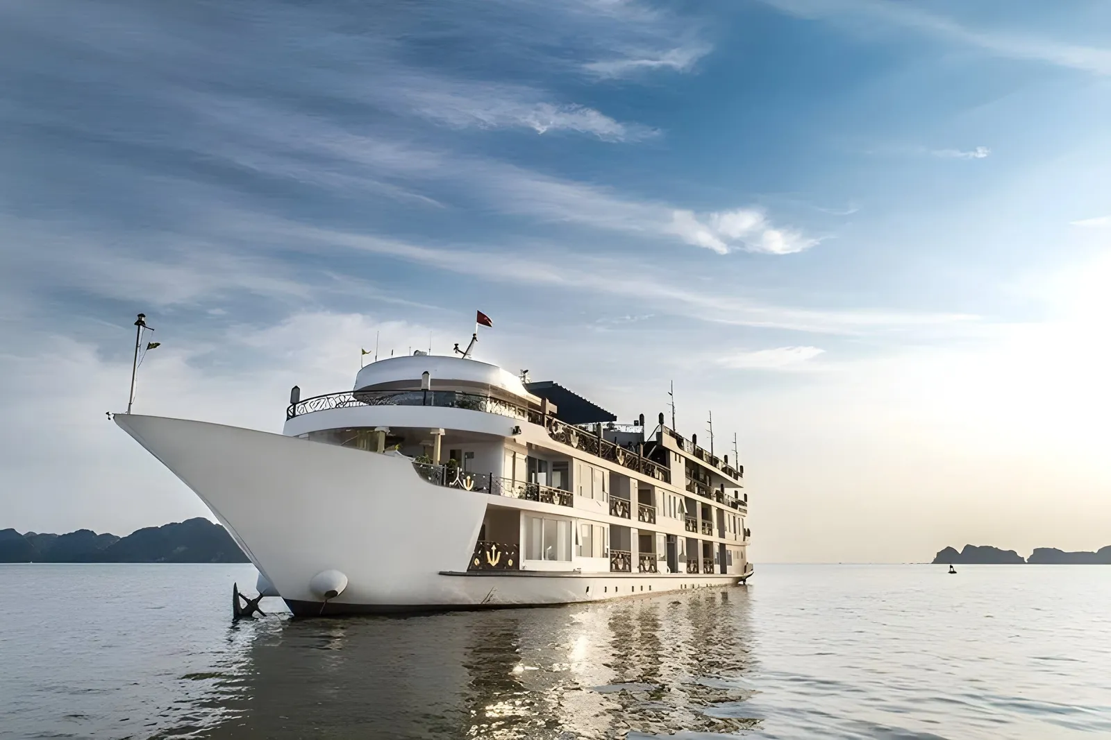 A large white multi-deck cruise ship with gold anchor emblems on its balconies sits on calm water, seen from the bow side, with distant limestone islands and a blue sky with wispy clouds behind it.