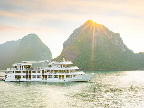 A white multi-deck cruise boat with 'ATHENA CRUISE' lettered on its side sails on calm green water in front of forested limestone karst peaks with the sun bursting over the summit.
