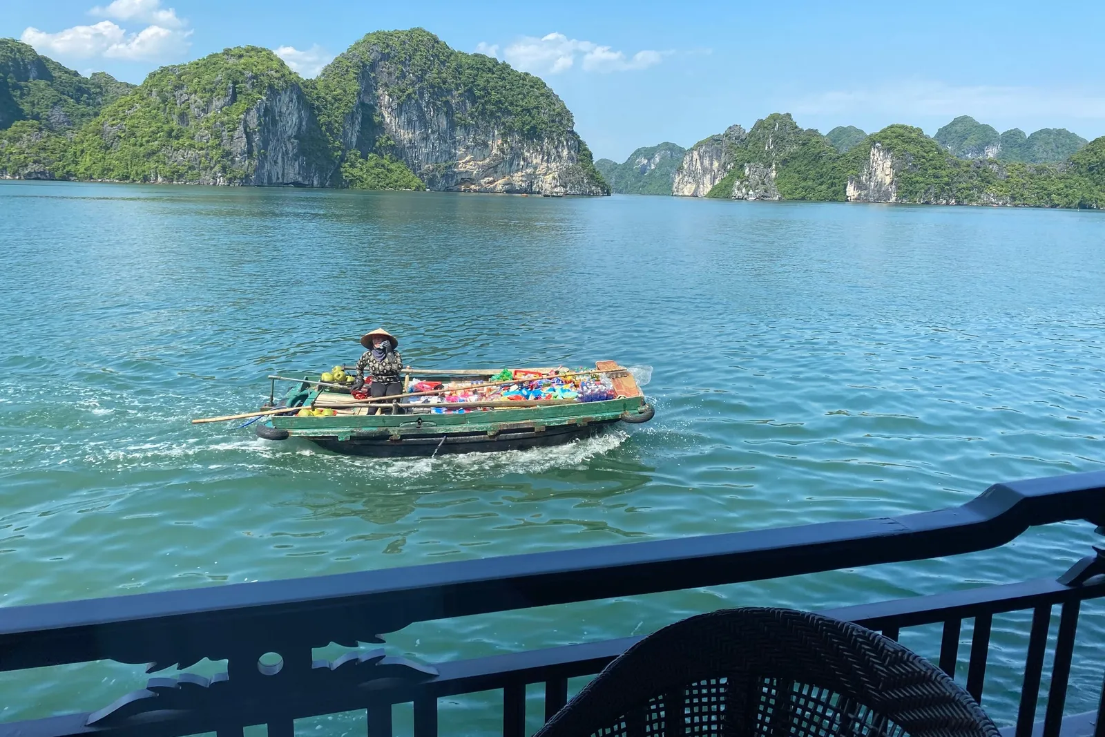 A vendor in a conical hat rows a small boat loaded with colorful goods across green bay water, seen from a dark railed balcony with a wicker chair in the foreground, with forested limestone karsts behind.
