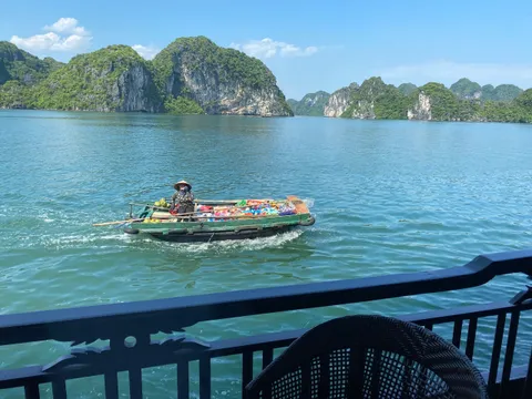 A vendor in a conical hat rows a small boat loaded with colorful goods across green bay water, seen from a dark railed balcony with a wicker chair in the foreground, with forested limestone karsts behind.