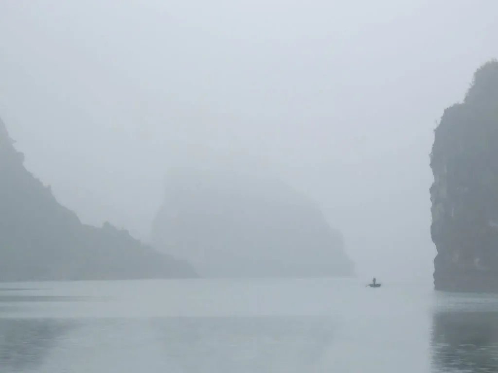 A small boat on grey water beneath misty limestone cliffs