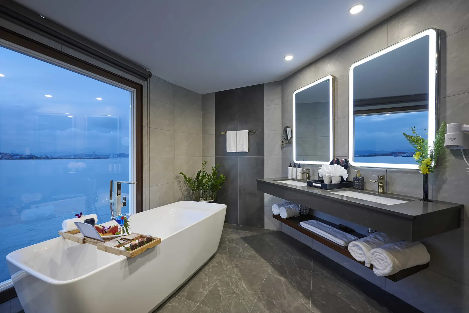 Suite bathroom with a freestanding tub beside a picture window, grey stone walls and twin backlit mirrors