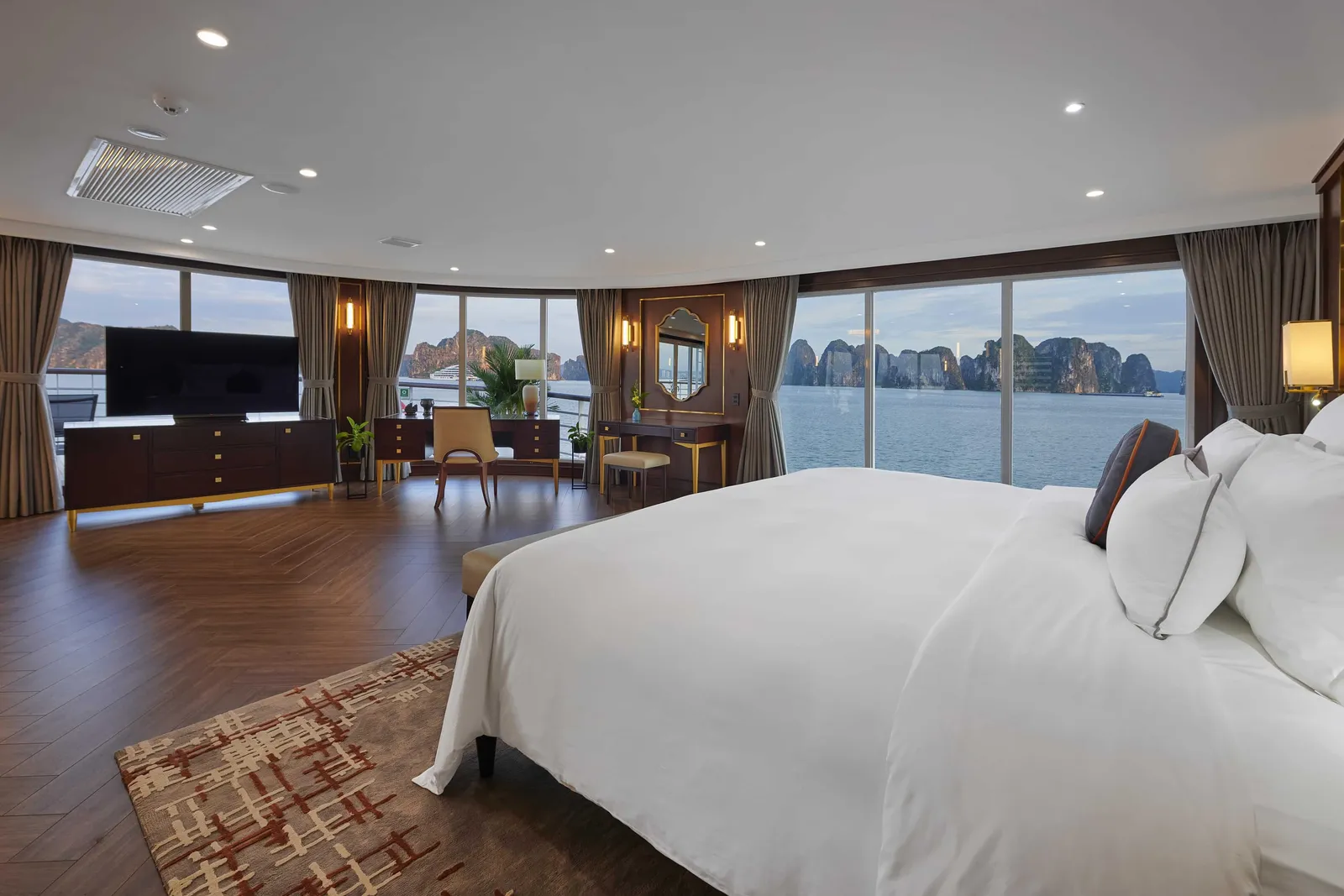 Suite bedroom with a herringbone timber floor, a wide bed and floor-to-ceiling windows wrapping round to karst views