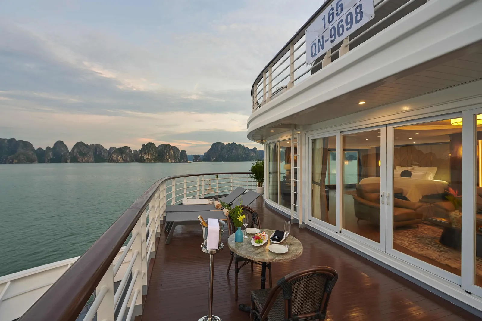 Wraparound suite terrace at the bow with loungers and a small table, the registration QN-9698 on the deck above, karst at dusk