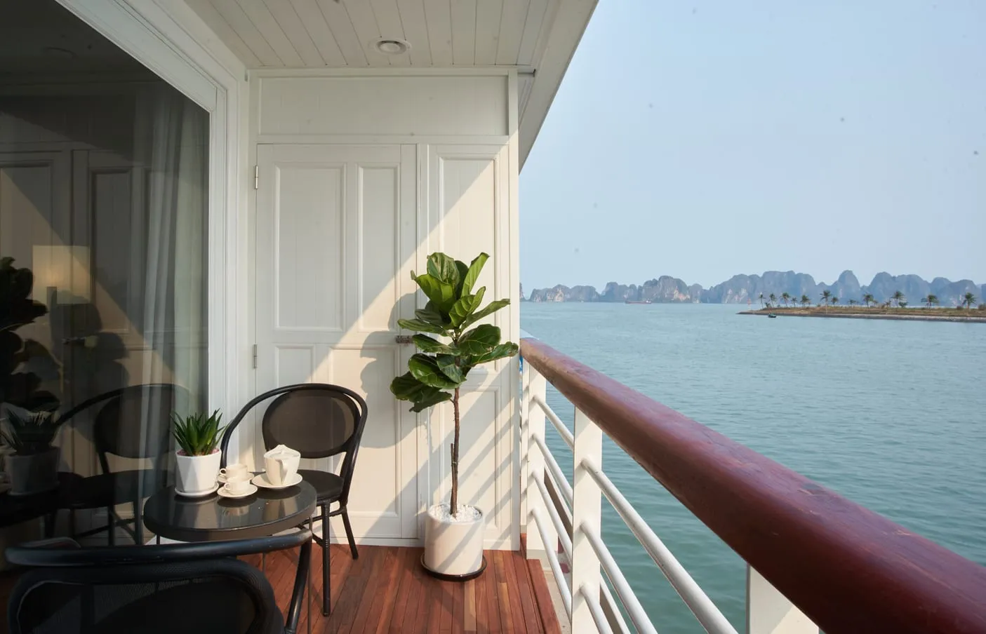 Private cabin balcony with two chairs, a tea set and a potted fig tree, the bay and distant karst beyond the rail