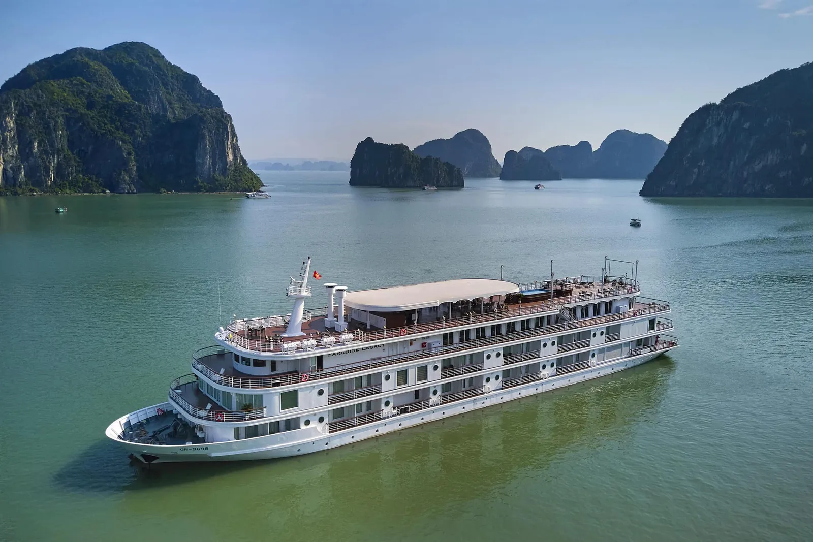 Paradise Legacy seen from above at anchor on green water, its sundeck canopy and stacked balcony decks in view, karst islands behind