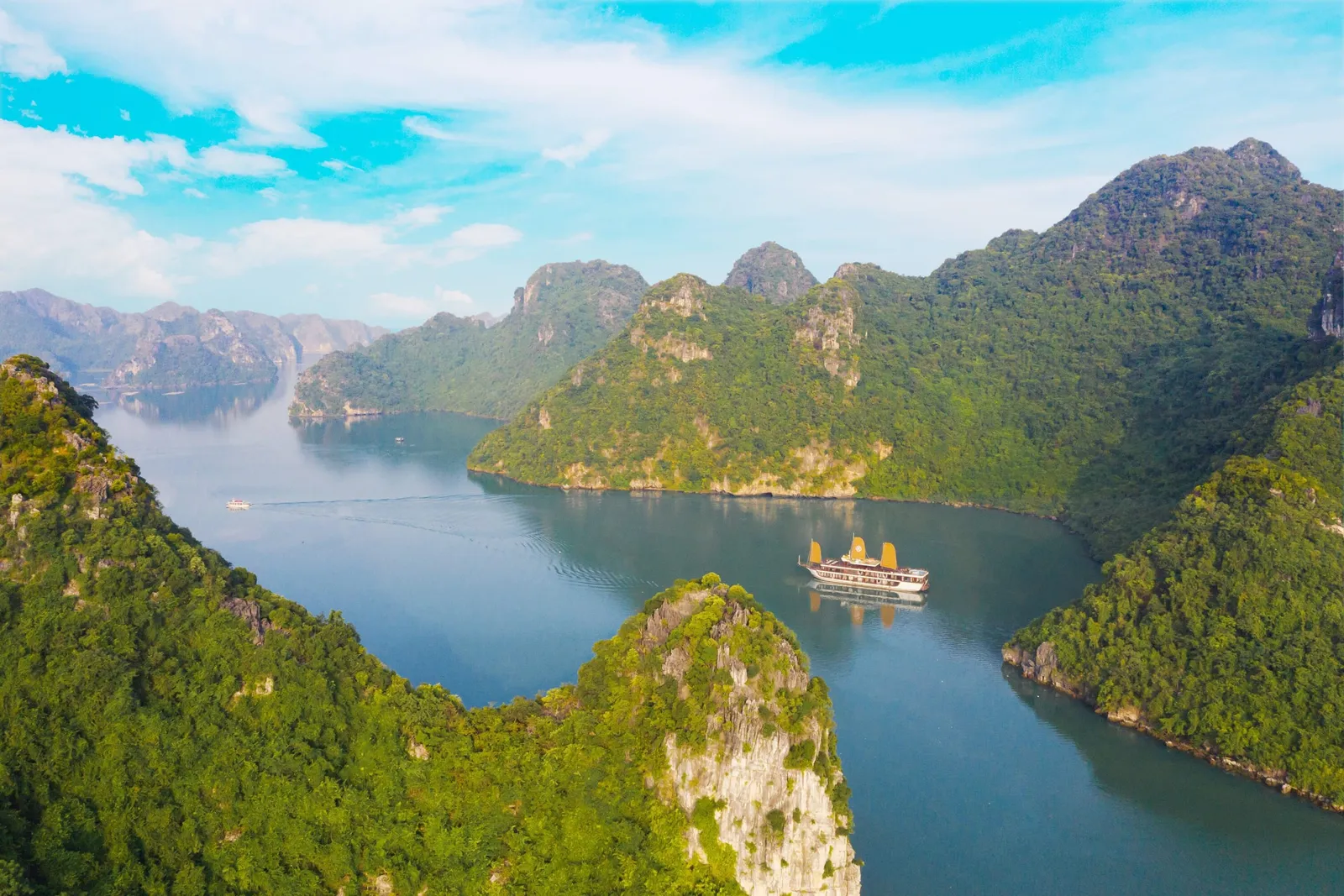 Aerial view of a cruise boat with golden-orange sails on calm blue-green water, surrounded by forested limestone karst peaks under a blue sky with clouds.