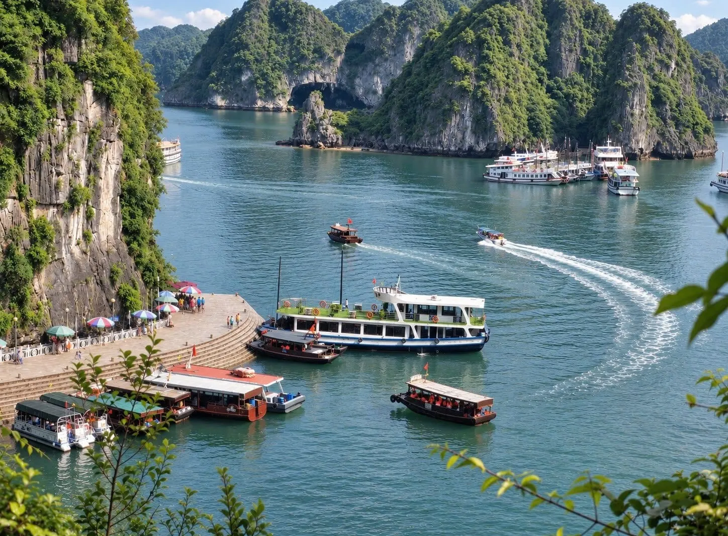 An elevated view of a green bay with limestone karst islands, tour boats and a ferry near a waterfront pier with umbrellas and people, a speedboat leaving a white wake, and foliage framing the foreground.