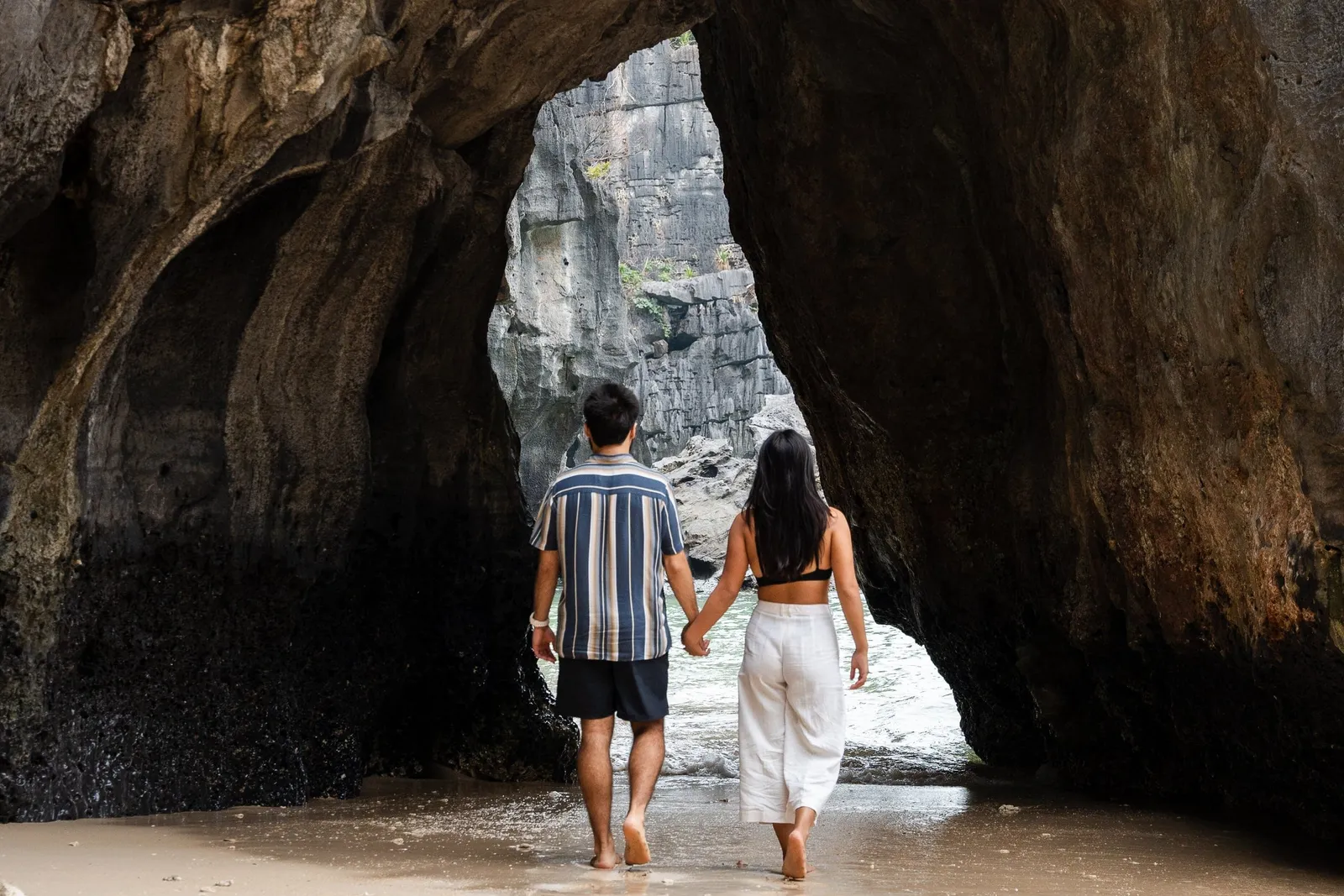 A couple holds hands and walks barefoot on a sandy beach toward a large limestone rock arch opening onto the water, seen from behind.
