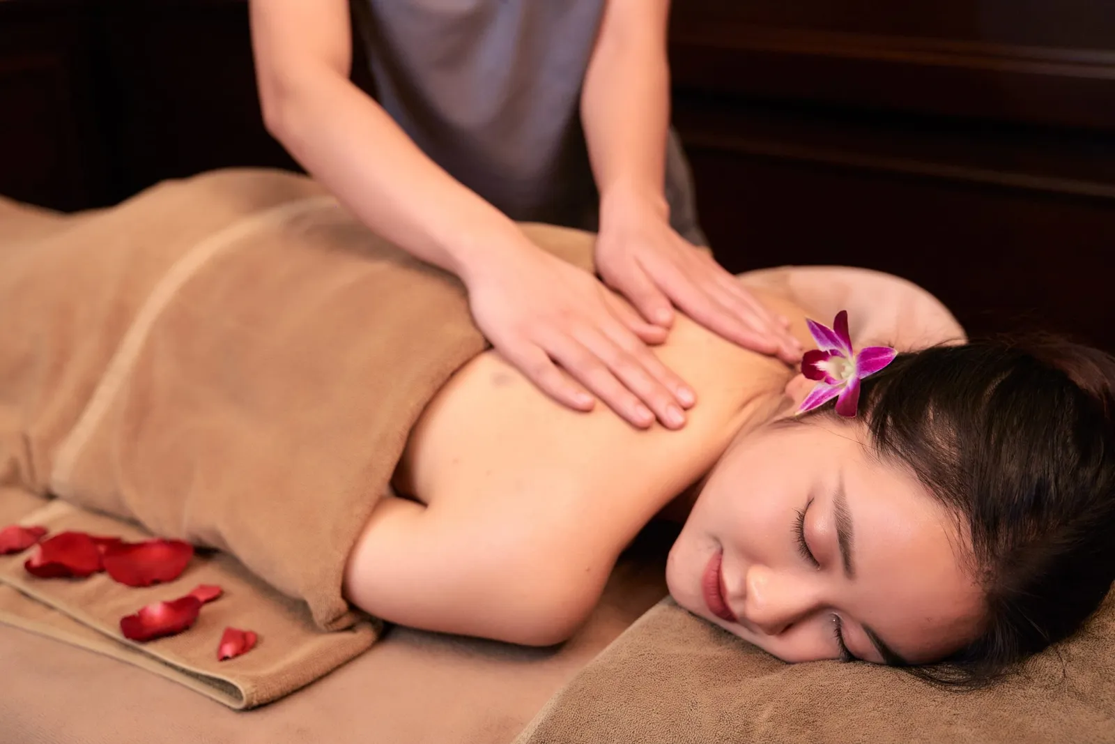 A woman lies face-down under a tan towel with an orchid in her hair and red rose petals nearby while a therapist's hands massage her upper back.
