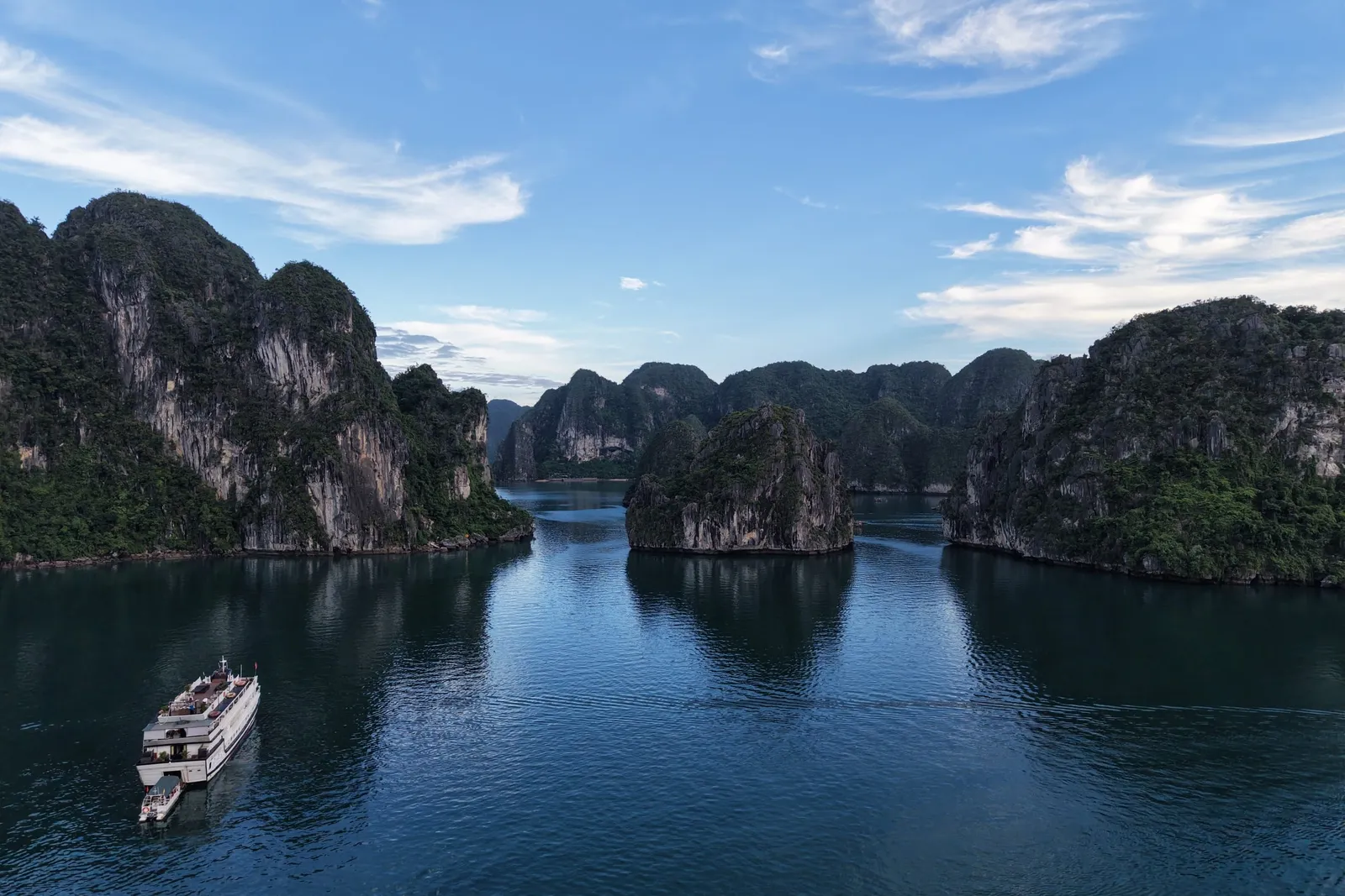 Aerial view of calm dark-blue water between forested limestone karst islands under a blue sky with wispy clouds, with a small white cruise boat and tender at lower left.