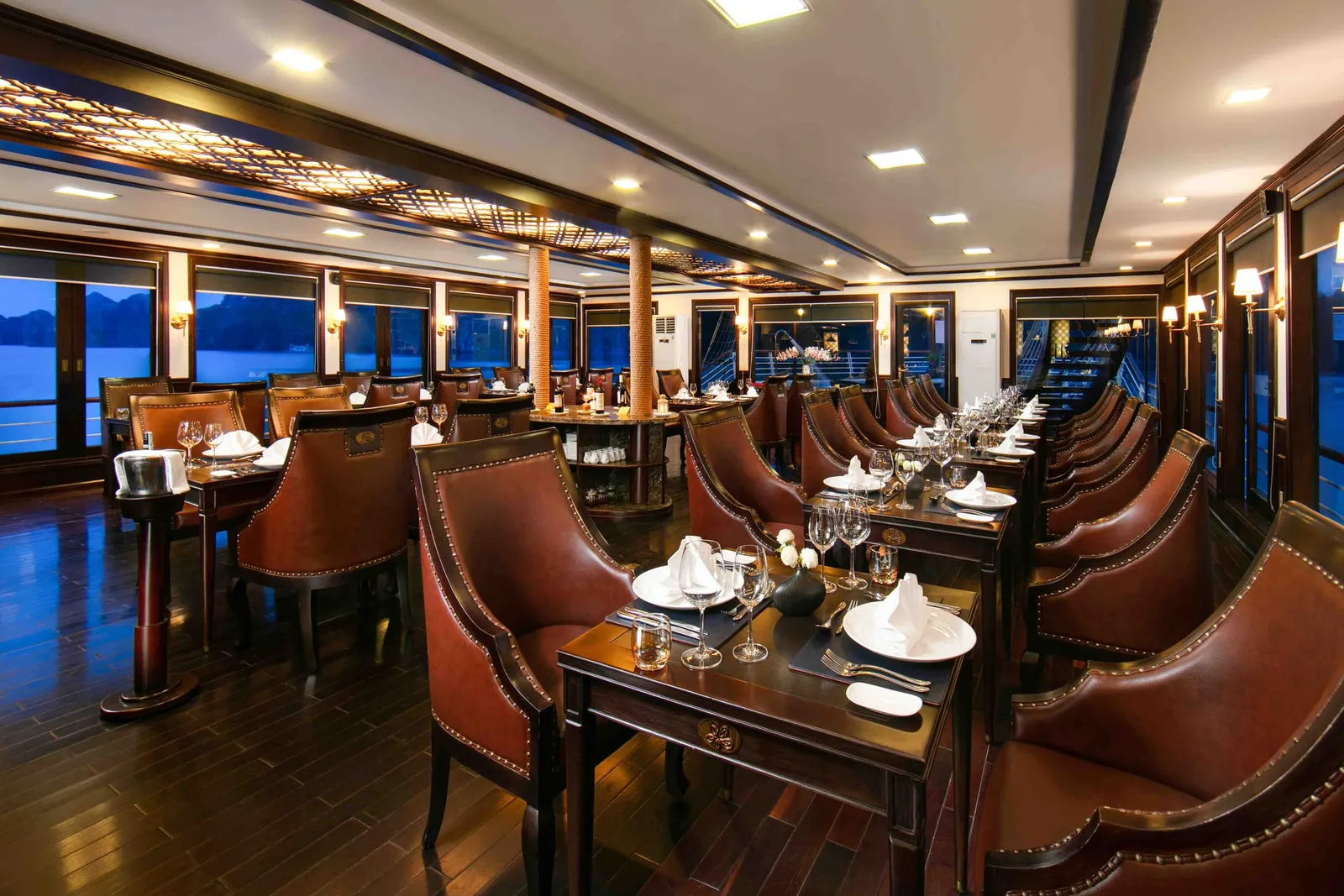 Interior of a cruise boat dining room with set tables, leather chairs, dark wood floors, and windows showing water and distant limestone karst silhouettes at dusk.
