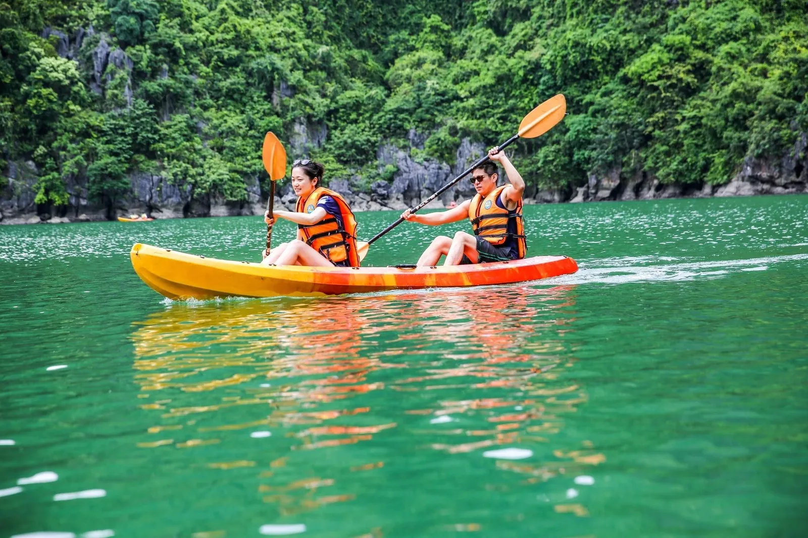 Two people in orange life vests paddle a yellow-orange tandem kayak on green water in front of a forested limestone cliff, with another kayak in the distance.