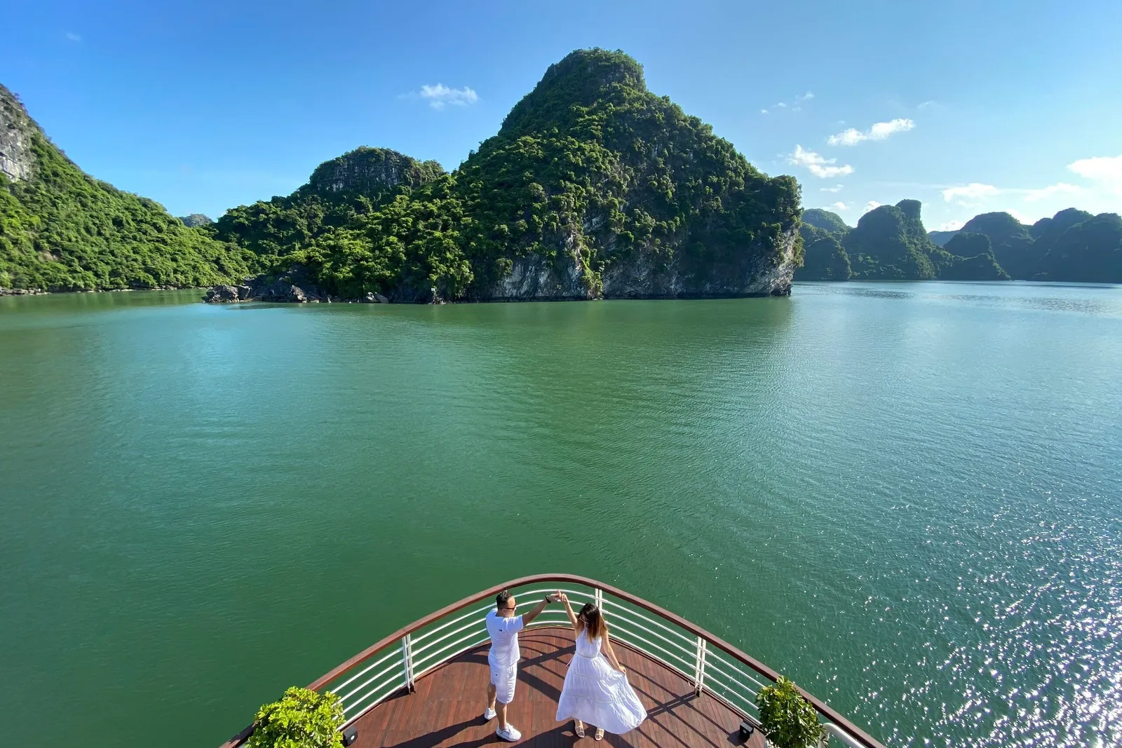 A couple in white dances on the bow deck of a boat, with green karst islands and calm green water of the bay ahead under a blue sky.