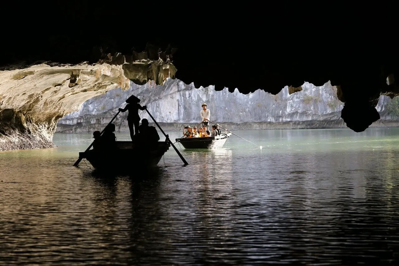 From inside a dark cave opening, two small rowing boats carry tourists and rowers, one in silhouette wearing a conical hat, on calm green water with limestone cliffs beyond.