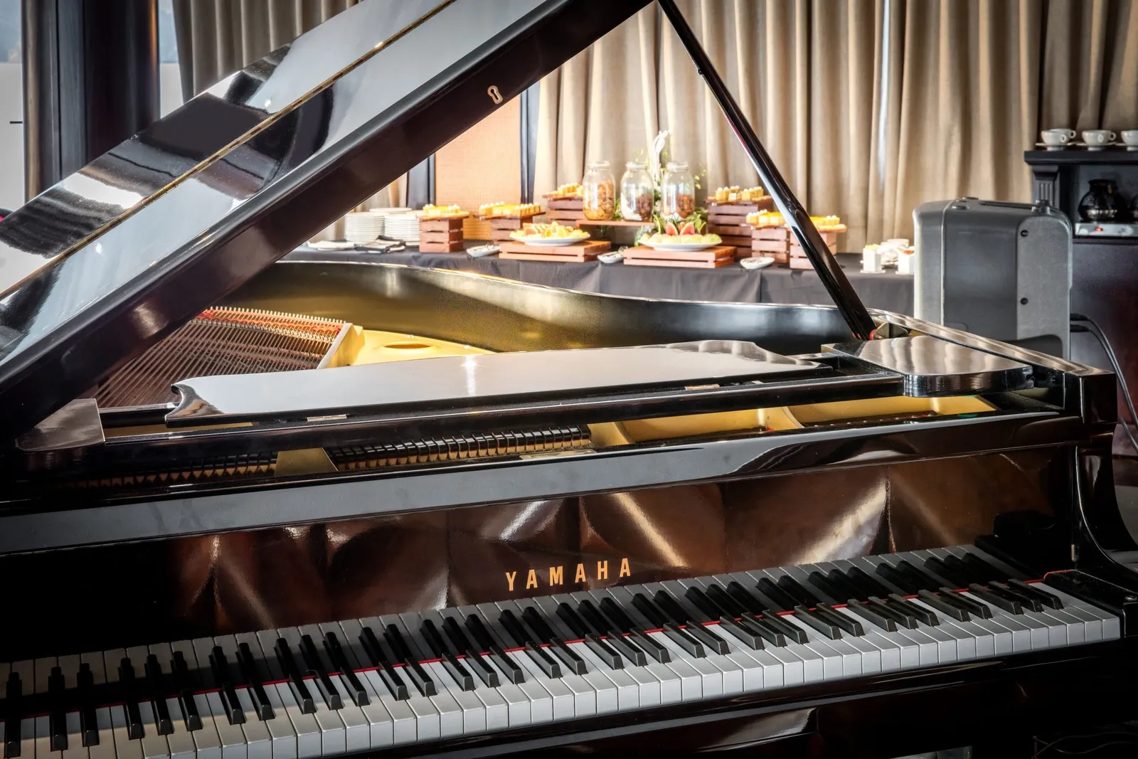 A black Yamaha grand piano with its lid raised stands in an indoor lounge, with a buffet of food displays, jars, plates and curtains behind it.