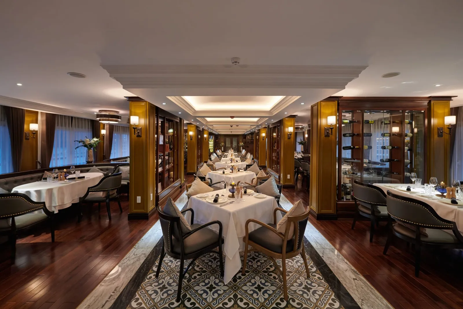 A long, elegant dining room with set tables with white tablecloths, upholstered chairs, wine display cabinets, wall sconces, wooden floors and a patterned tile runner down the center, with curtained windows on the left.