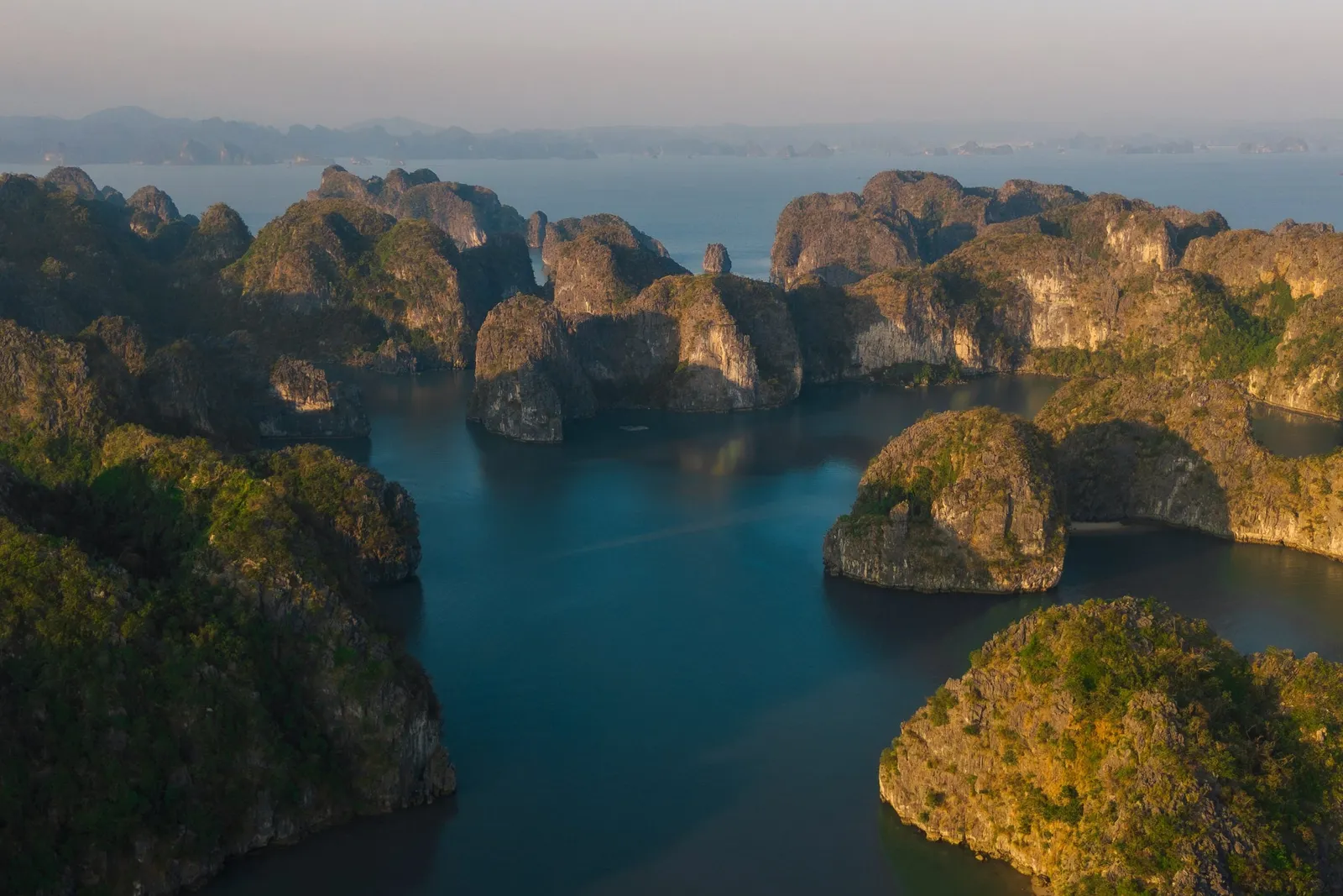 An aerial view at golden hour of dark blue water surrounded by forested limestone karst islands, with hazy hills in the distance.