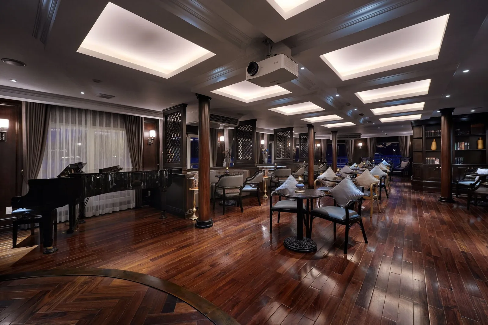 A dark-wood lounge with a grand piano, polished hardwood floor, small round tables with cushioned chairs, lit ceiling panels and a ceiling-mounted projector.