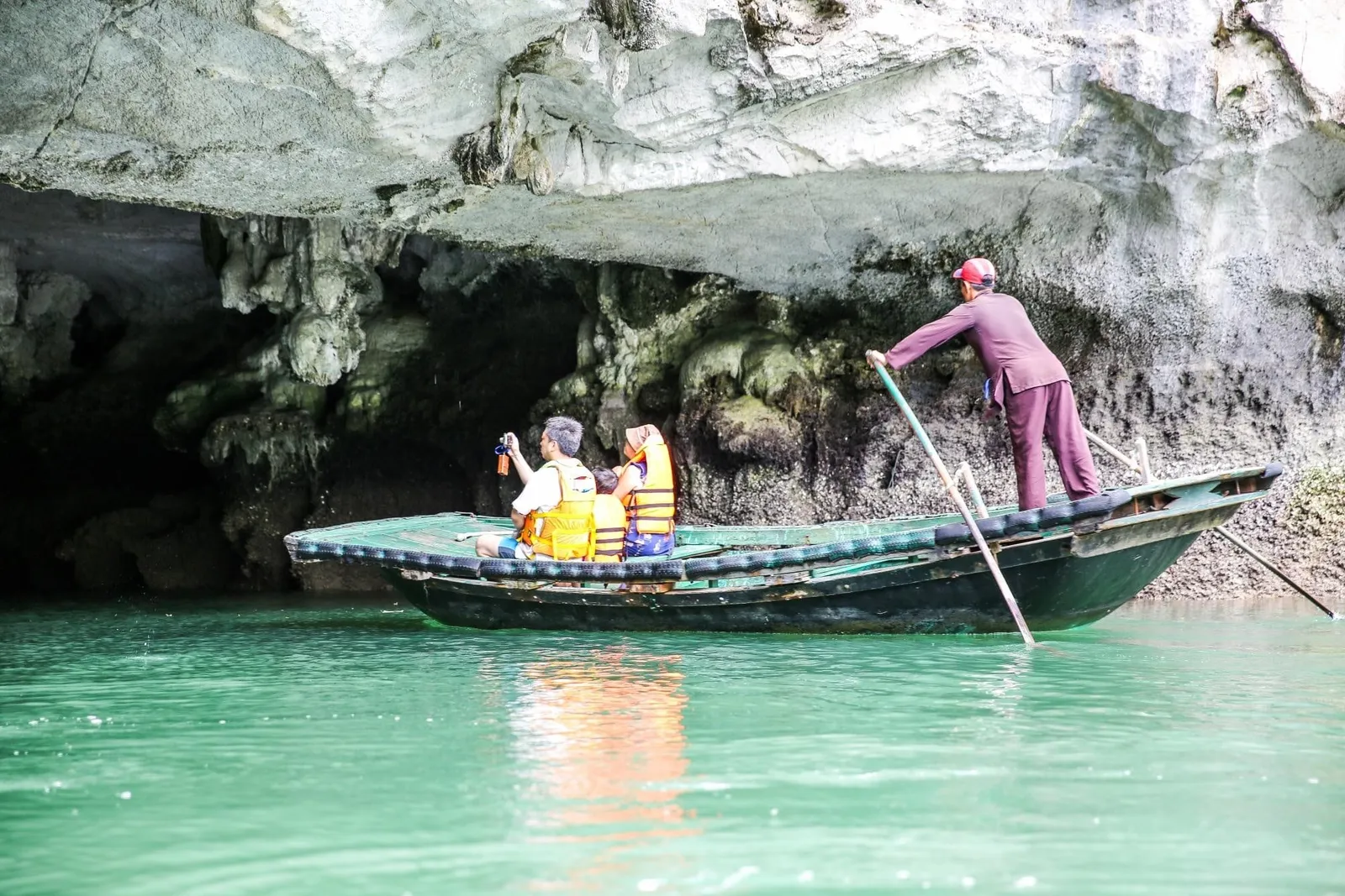 A rower stands in a small green rowing boat carrying tourists in orange life jackets in front of a limestone cave overhang above green water.