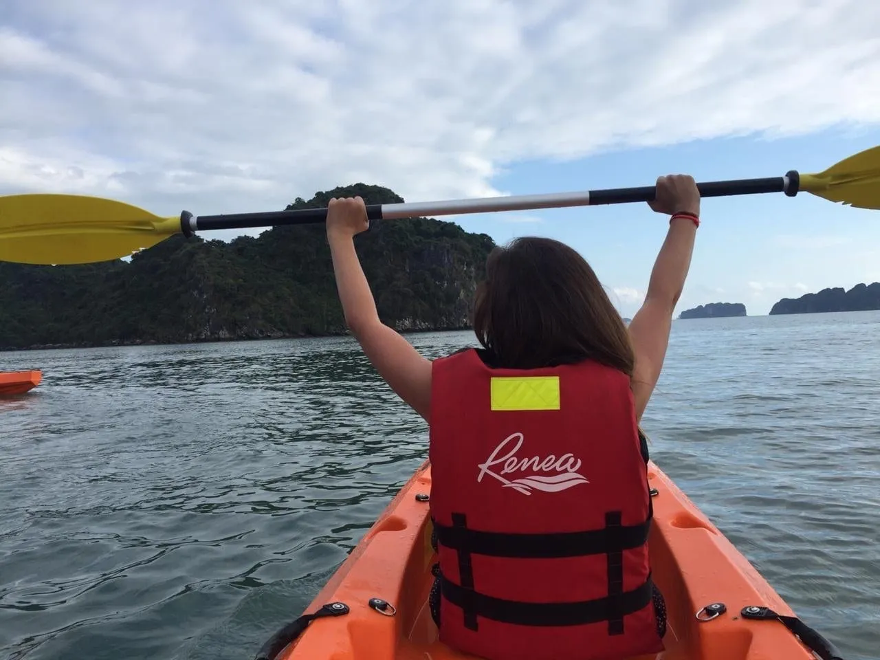 A person in a red life vest printed with 'Renea' sits in an orange kayak holding a yellow paddle overhead, facing calm water and forested limestone islands under a cloudy sky.