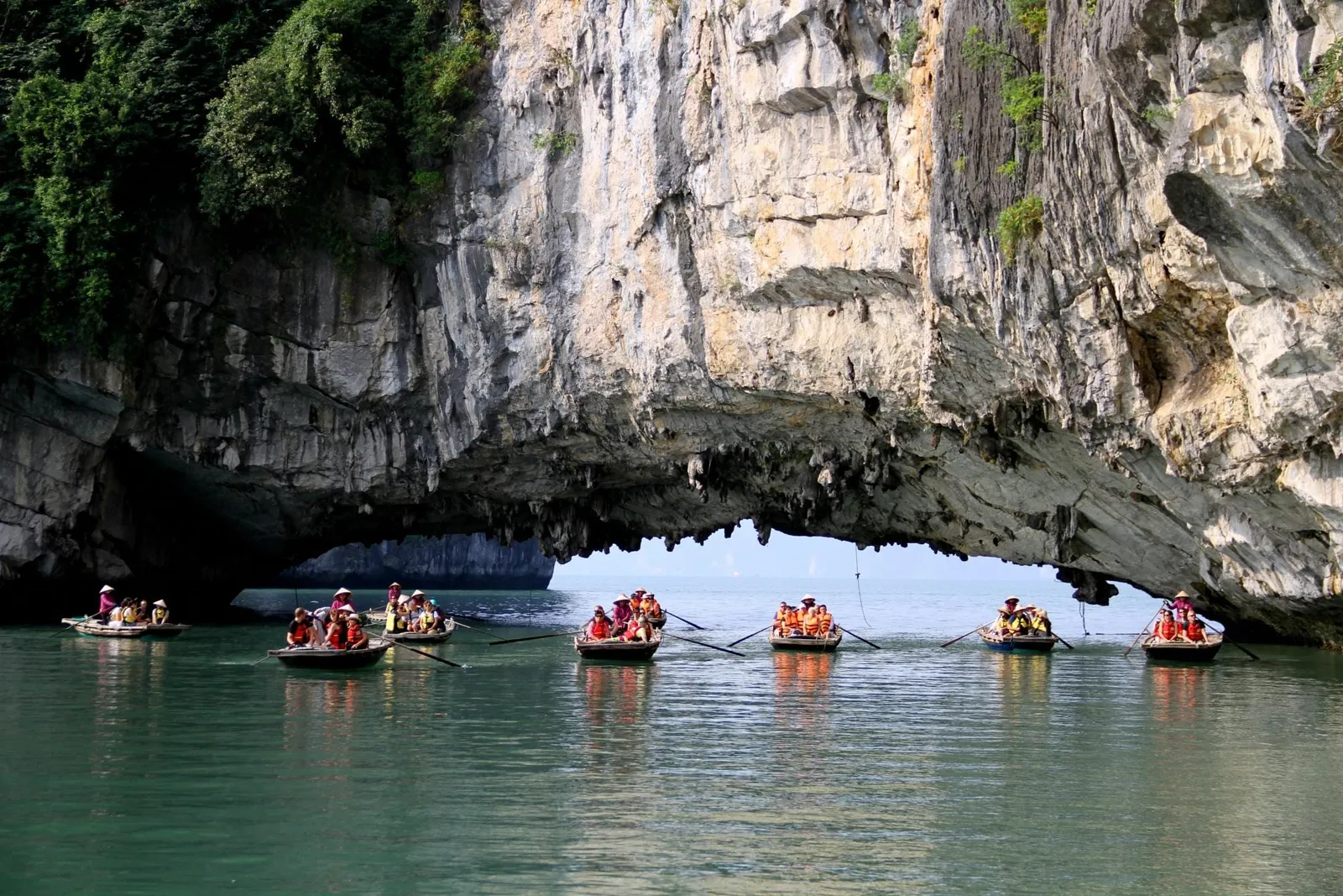Several small rowing boats carrying tourists in orange life jackets and conical hats float on green water in front of a large limestone cliff arch.