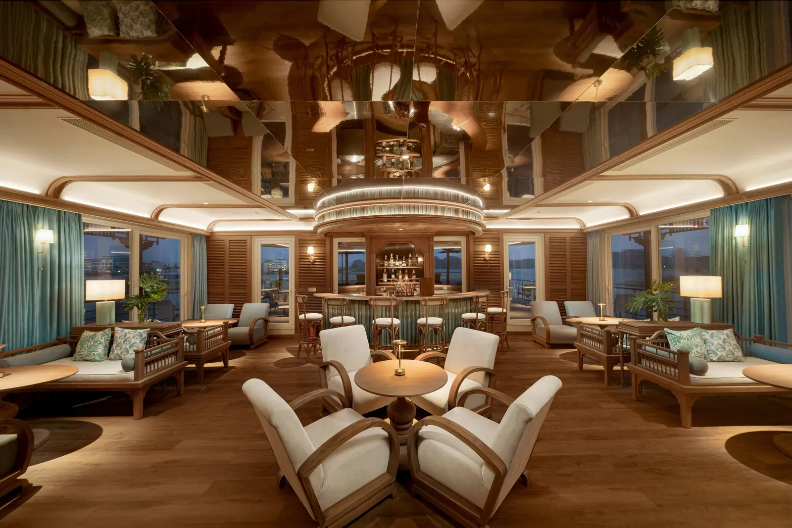 A lounge with a central round bar, bar stools, wooden armchairs, sofas with patterned cushions, teal curtains and a mirrored ceiling, with dusk light visible through the windows.