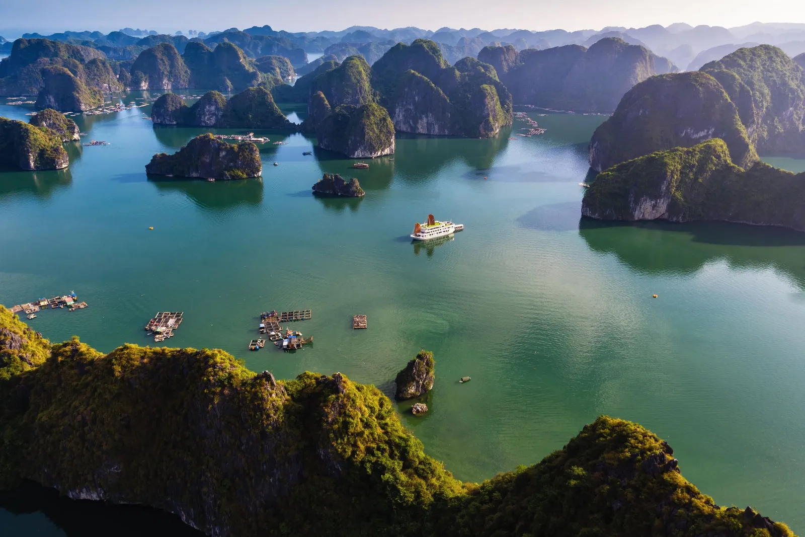 Aerial view of green water dotted with forested limestone karst islands, with a single white cruise boat with orange sails and a few floating fish farms in the middle distance.