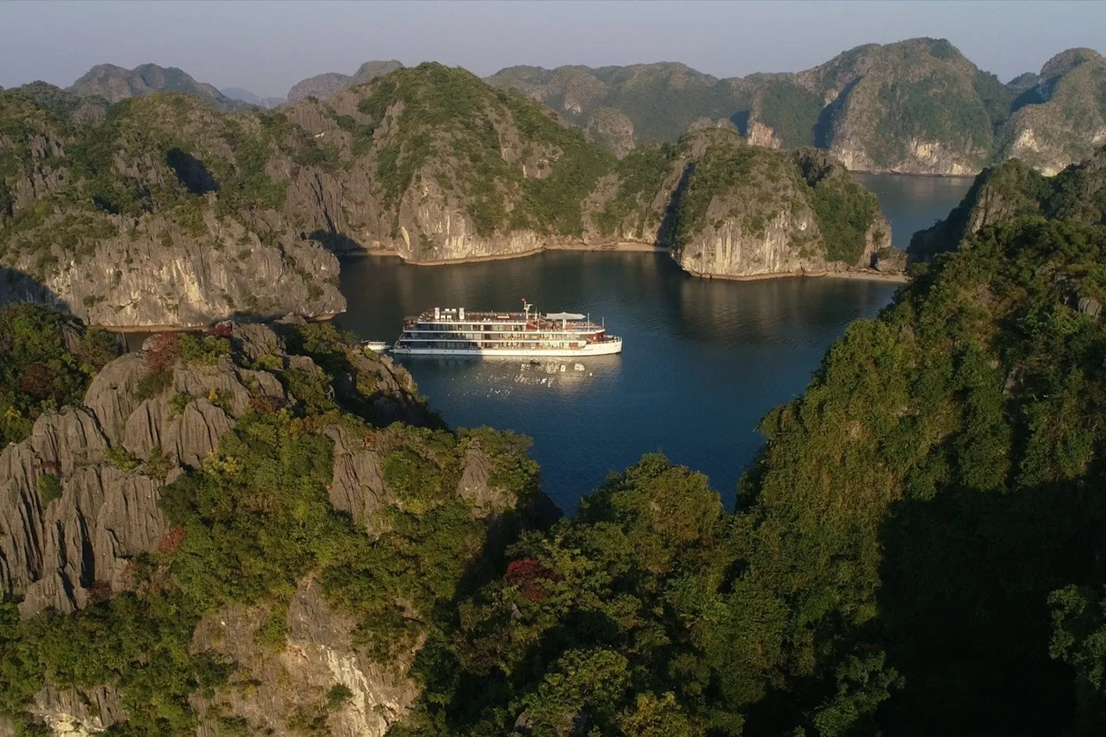 Aerial view of a white multi-deck cruise ship anchored in a sheltered blue bay surrounded by forested limestone karst hills.