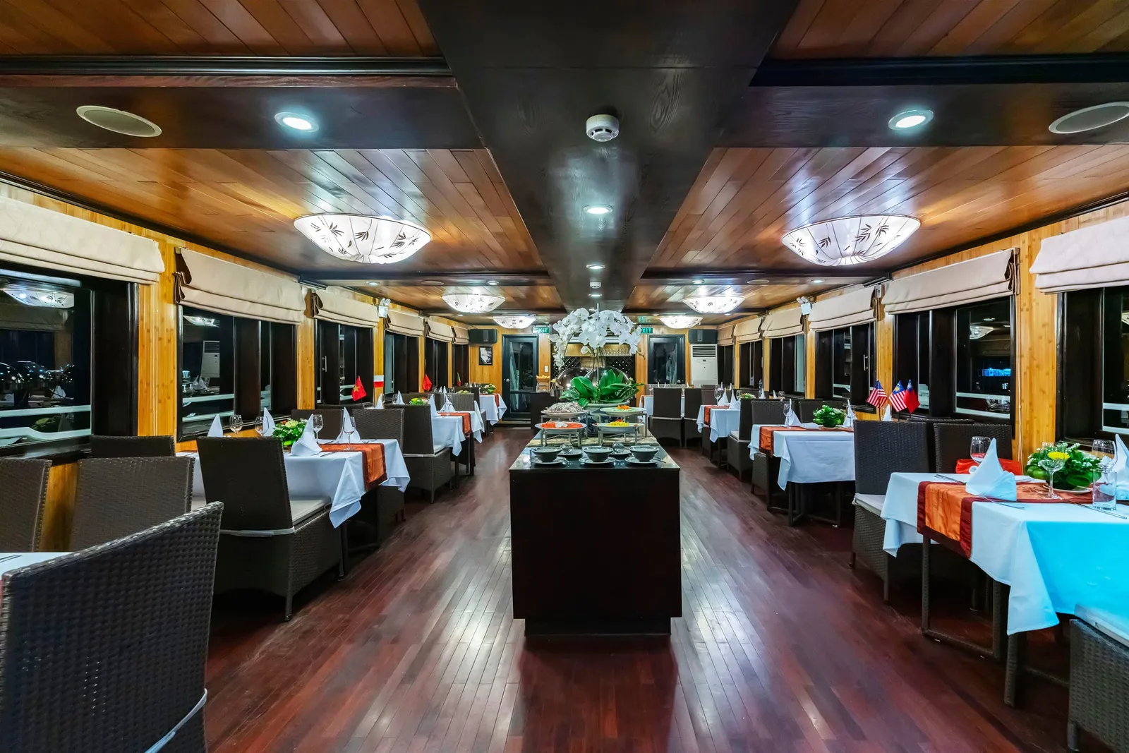 Interior of a wood-paneled dining room with set tables, wicker chairs, a central buffet counter with white orchids, and small Vietnamese and American flags on the tables, with dark windows on both sides.