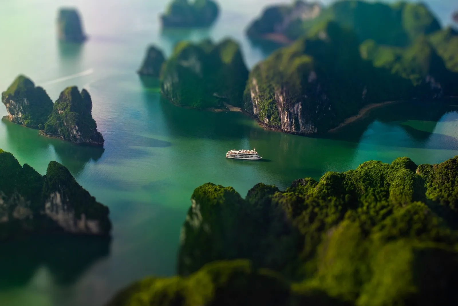 An elevated aerial view with a tilt-shift effect shows a small white cruise boat on green water among forested limestone karst islands.