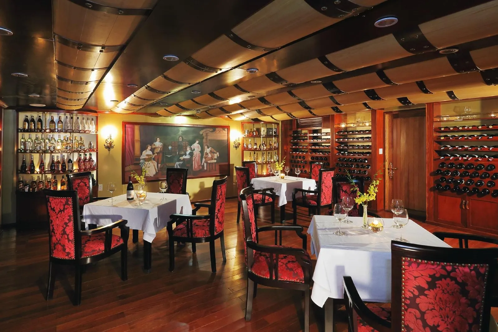 An indoor restaurant with white-clothed tables, red damask-patterned wooden chairs, wine racks and bottle shelves, a framed painting, and a barrel-stave style ceiling over a dark wood floor.