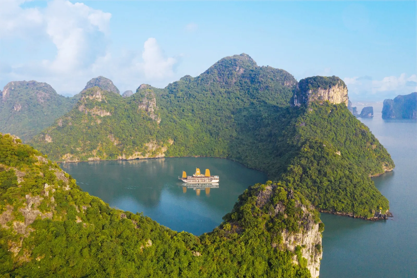 Aerial view of a small cruise boat with golden-orange sails on a calm green-blue lagoon enclosed by forested limestone karst hills under a blue sky with clouds.