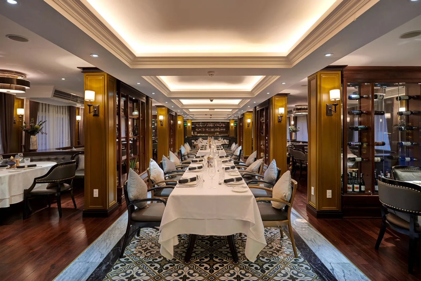 A long elegant dining room with a white-clothed table set with napkins and glasses, cushioned chairs, gold-toned wood pillars, a wine display cabinet, and a patterned tile floor inset.
