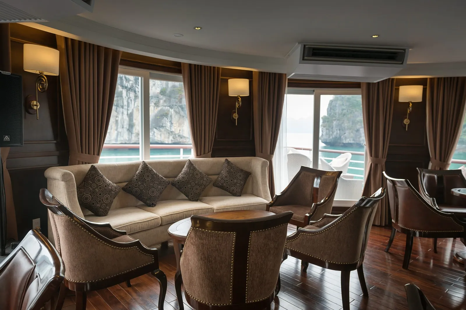 A lounge interior on a boat with a cream sofa, upholstered armchairs, wall lamps and brown curtains, with windows showing limestone karst cliffs and green water outside.