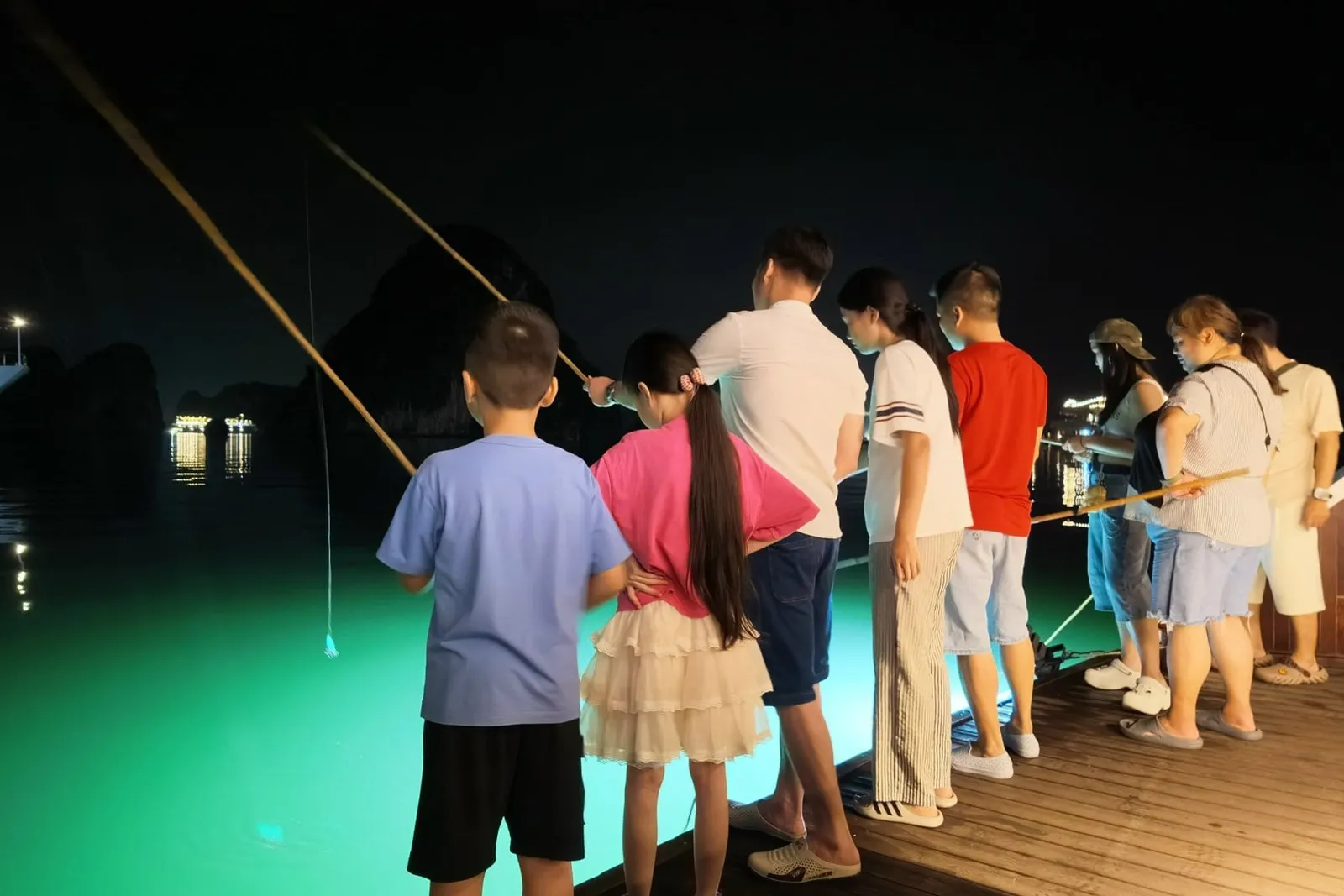 At night, a group of children and adults stand on a wooden deck holding long squid-fishing rods over glowing green water, with a lit ship bearing painted lettering on its hull and dark limestone karsts in the background.