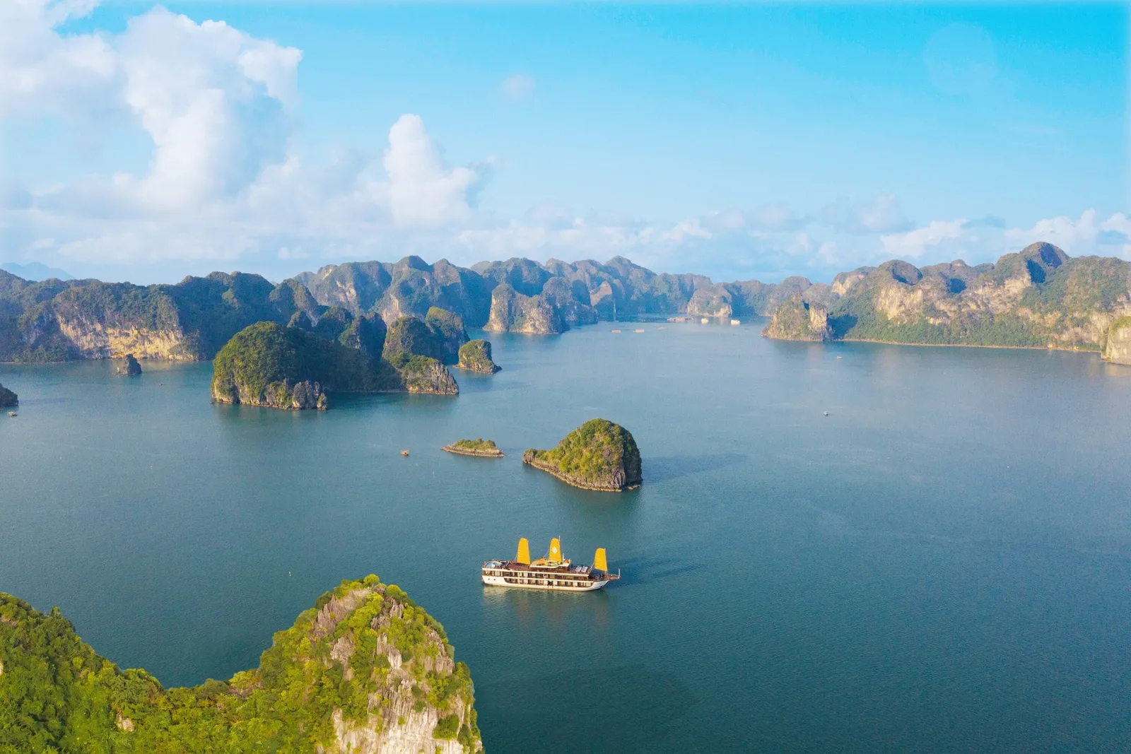 Aerial view of a cruise boat with yellow sails on calm green-blue water, surrounded by forested limestone karst islands under a blue sky with clouds.