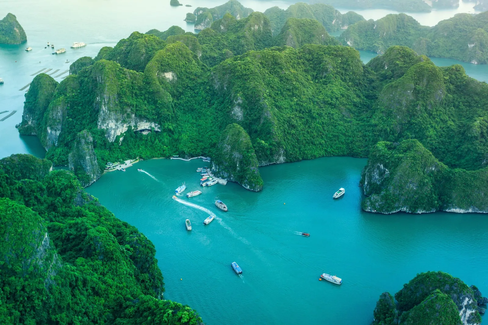 Aerial view of turquoise water enclosed by green forested limestone karst islands, with several small tour boats and wakes near a landing area.