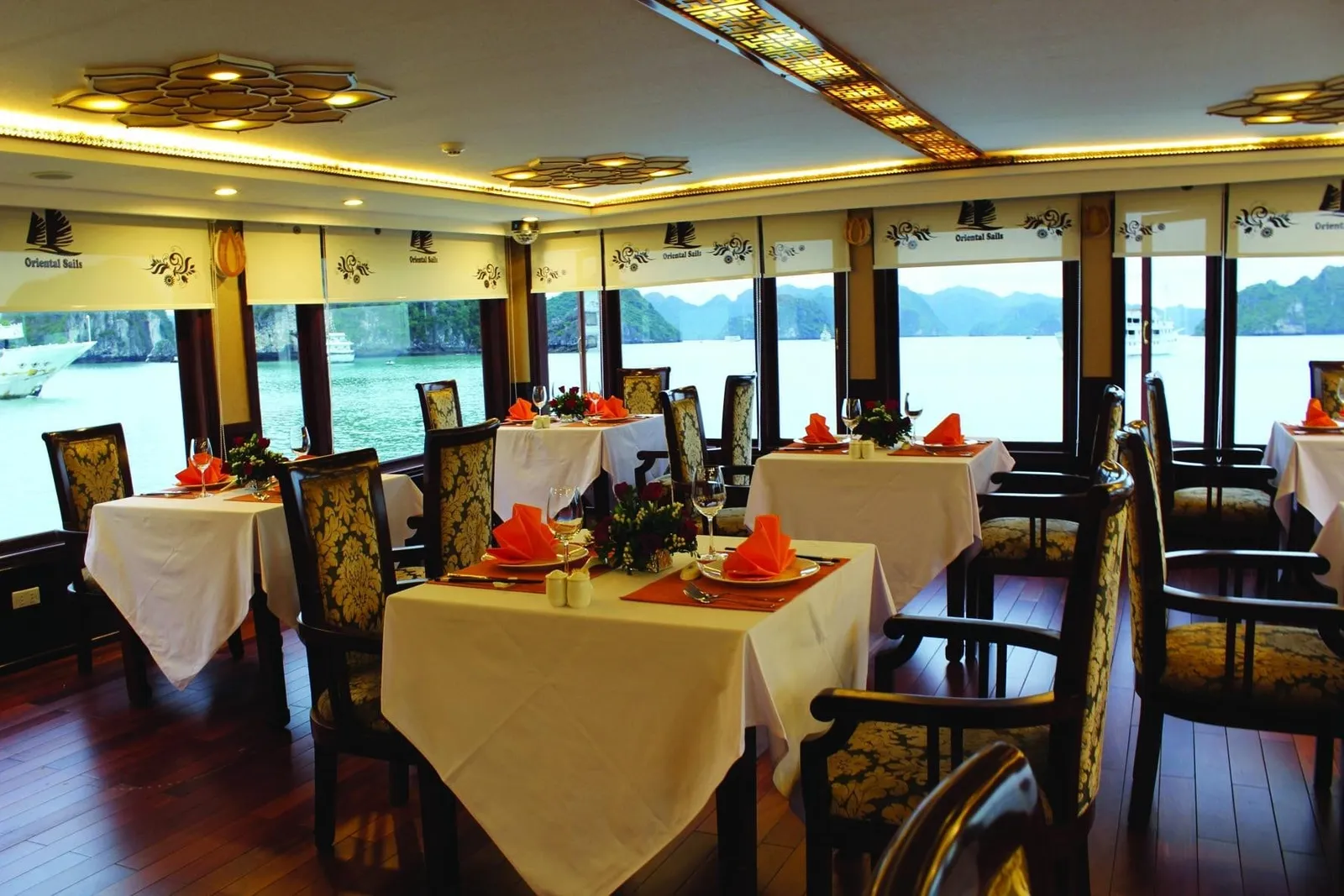 Interior dining room on a boat with tables set with white cloths, orange napkins and flowers, with window blinds printed 'Oriental Sails' and limestone karsts and other vessels visible outside.