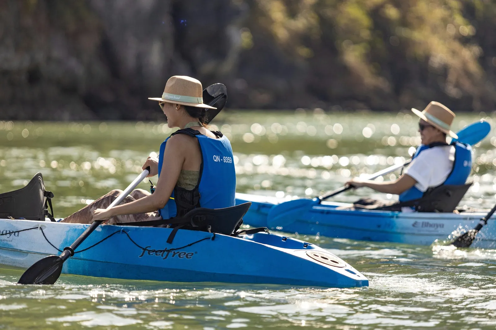 Two people in straw hats and blue life vests paddle blue kayaks marked 'feelfree' and 'Bhaya' on green water, with a blurred wooded cliff behind them.