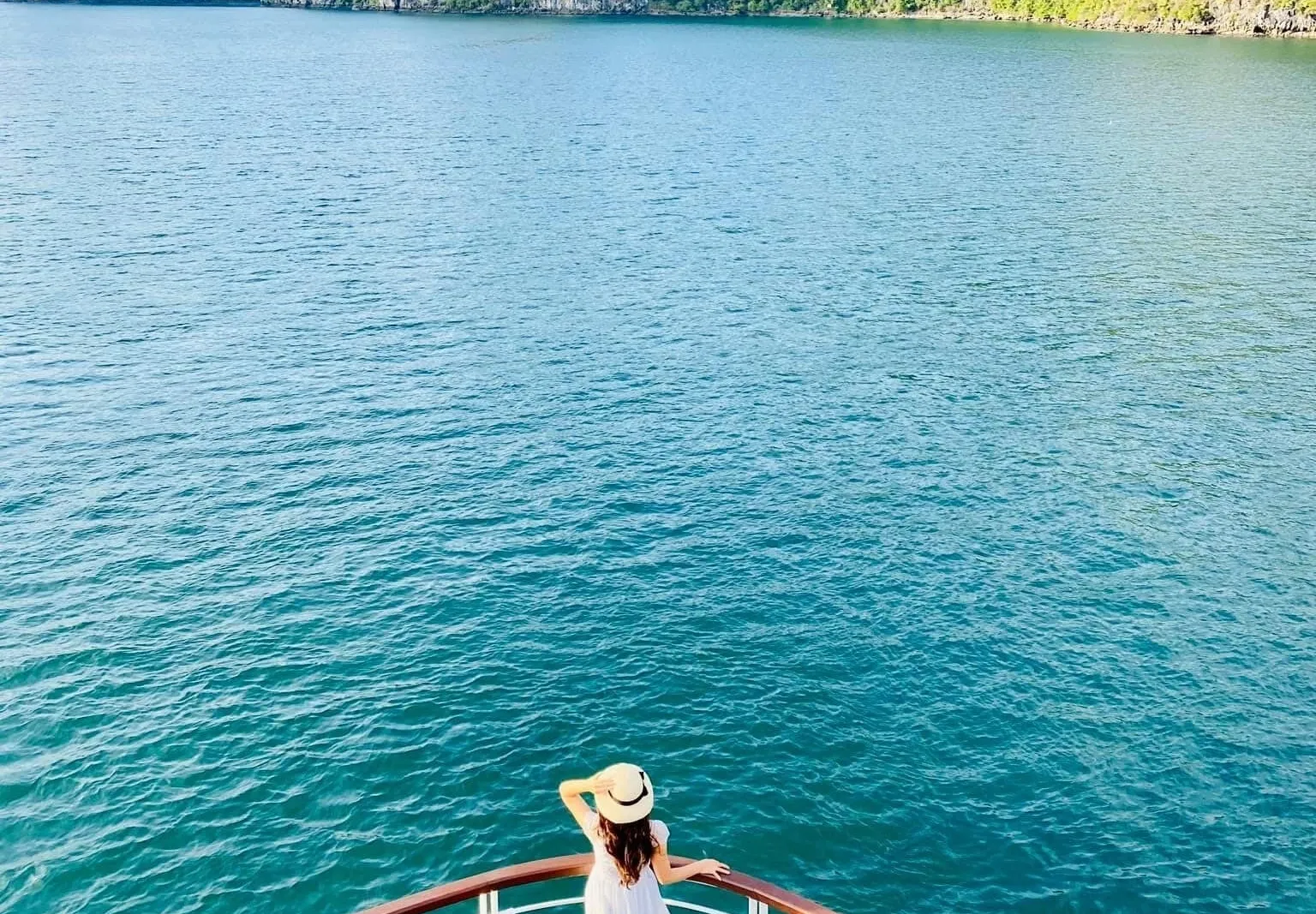 A woman in a white dress and straw hat stands at the curved wooden bow deck railing of a boat, looking out over turquoise water toward green limestone karst islands.